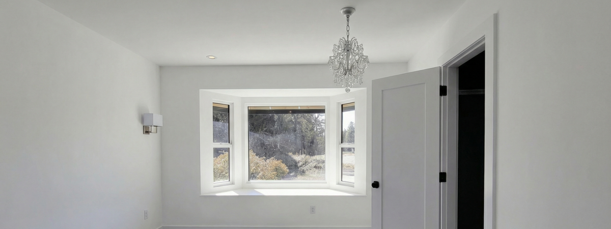 A fully renovated, brightly lit room painted in crisp white. The focal point is a newly installed bay window with a deep built-in window seat. The space features a decorative chandelier, a modern wall sconce, and a paneled white interior door with matte black hardware by PCC.