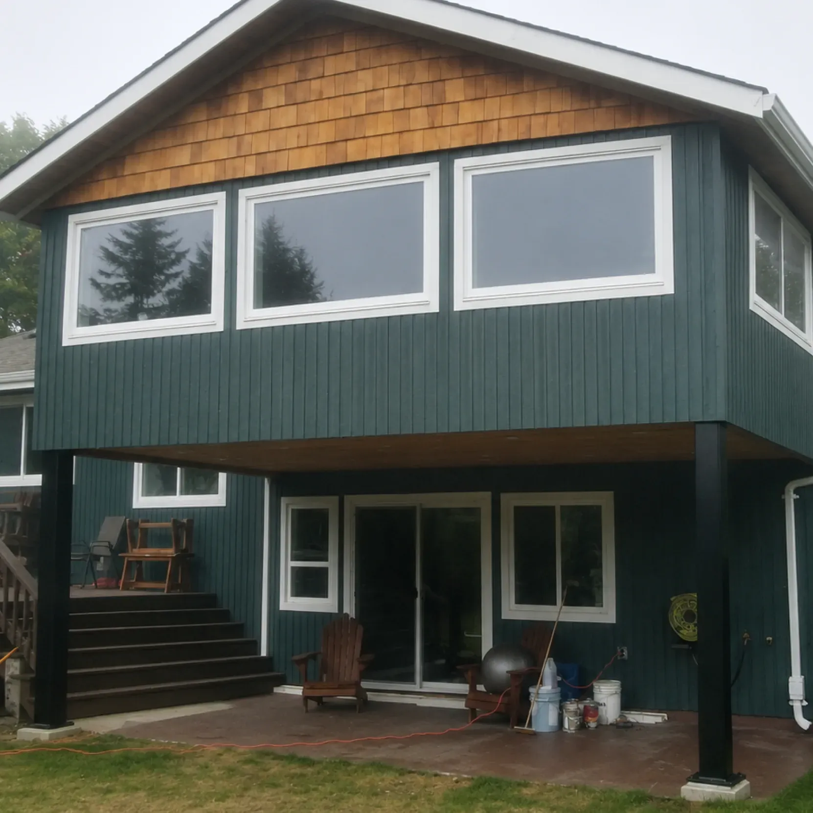 Completed second-story home extension featuring dark green exterior siding, natural wood shingle gable accents, and newly installed large windows over a concrete patio by PCC Renovations.
