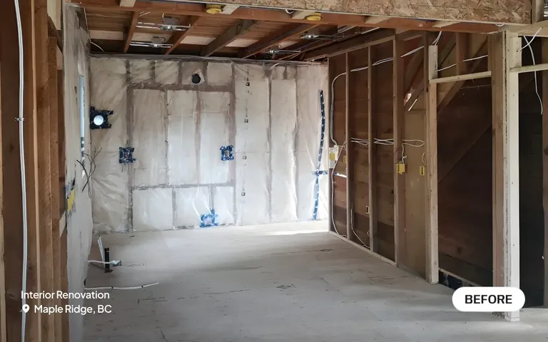 A before photo of an interior renovation in Maple Ridge, BC, showing a space completely stripped down to the wooden wall studs and ceiling joists, with exposed insulation and rough-in electrical wiring by PCC Renovations. 