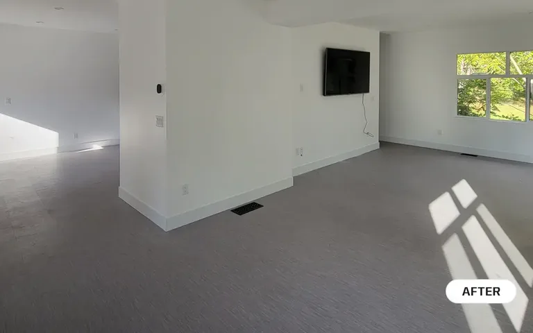 An after photo of a bright, newly renovated living space featuring light grey flooring, smooth white walls, new baseboards, and a wall-mounted television by PCC Renovations.