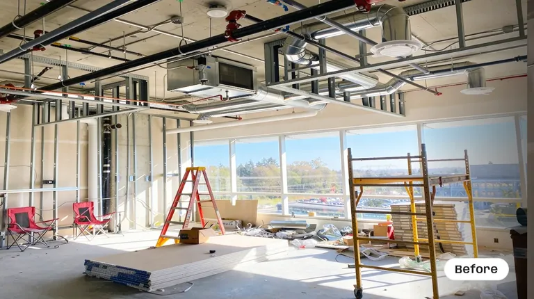 A commercial office space during the early construction phase, featuring a raw concrete ceiling with exposed HVAC ductwork and red fire suppression pipes. The room shows bare metal wall framing, large exterior windows, yellow scaffolding, a red step ladder, and stacks of drywall on the floor by Pacific Coast Contracting Renovations.