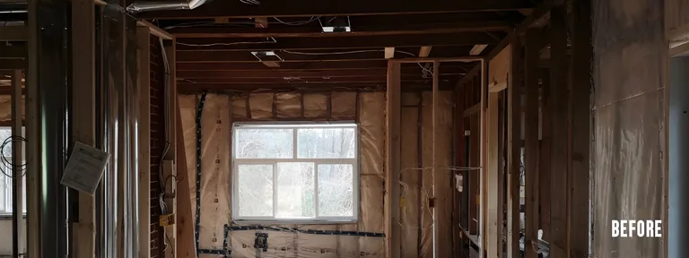 A room interior undergoing a major renovation, stripped completely down to the wooden wall studs and ceiling joists. The space features exposed insulation behind a plastic vapor barrier, rough electrical wiring, metal ductwork, and a standard rectangular window.