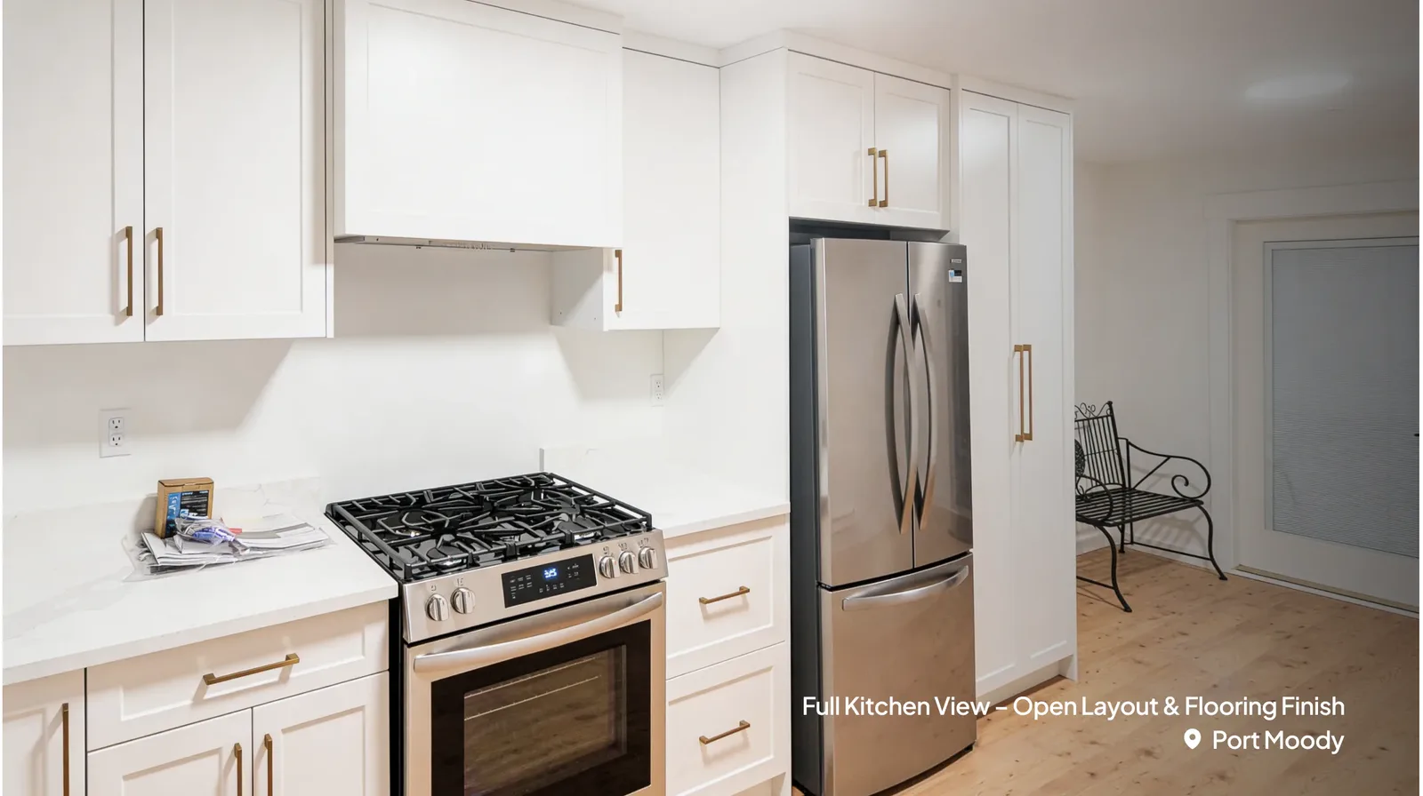 A close-up view of a newly renovated kitchen featuring white shaker cabinets with gold hardware, a stainless steel gas stove, and a matching French-door refrigerator. Light wood flooring extends toward a bright entryway with a black wire bench by Pacific Coast Contracting Renovations.