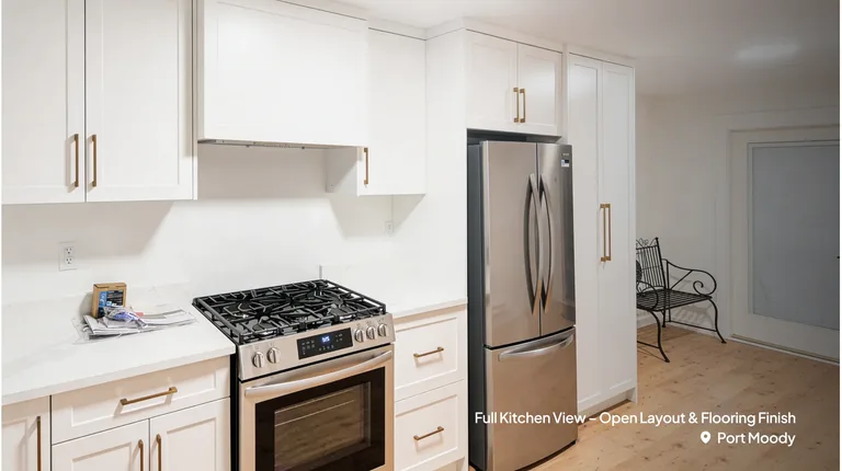 A close-up view of a newly renovated kitchen featuring white shaker cabinets with gold hardware, a stainless steel gas stove, and a matching French-door refrigerator. Light wood flooring extends toward a bright entryway with a black wire bench by Pacific Coast Contracting Renovations.
