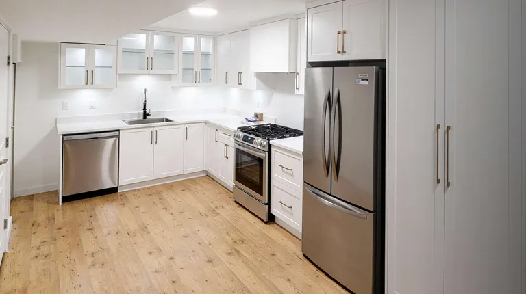 A wide shot of a modern, white L-shaped kitchen with stainless steel appliances, gold cabinet hardware and light wood floors in a basement secondary suite in Port Moody, Tri-Cities, by PCC Renovations.