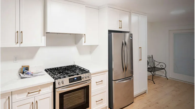 A close-up view of a newly renovated kitchen in a Port Moody basement suite featuring white shaker cabinets with gold hardware, a stainless steel gas stove, and a matching French-door refrigerator. Light wood flooring extends toward a bright entryway with a black wire bench by Pacific Coast Contracting Renovations.