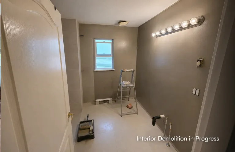 A bathroom in the early stages of renovation with grey walls, exposed plumbing pipes, a stepladder, and a paint tray on the floor. Text overlay reads: "Interior Demolition in Progress by PCC.