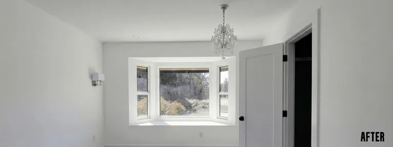 A fully renovated, brightly lit room painted in crisp white. The focal point is a newly installed bay window with a deep built-in window seat. The space features a decorative chandelier, a modern wall sconce, and a paneled white interior door with matte black hardware by PCC Renovations.