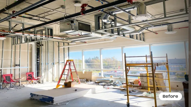 A commercial office space during the early construction phase, featuring a raw concrete ceiling with exposed HVAC ductwork and red fire suppression pipes. The room shows bare metal wall framing, large exterior windows, yellow scaffolding, a red step ladder, and stacks of drywall on the floor by Pacific Coast Contracting Renovations. — before
