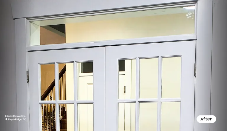 A close-up view of newly installed, white multi-paned French doors featuring dark hardware and a rectangular glass transom window above. Through the glass panels, a staircase with a wooden handrail and a brightly lit hallway are visible.