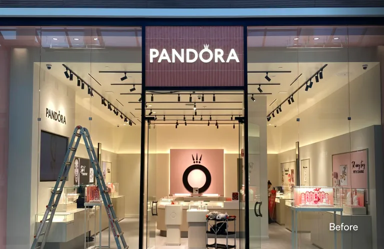 A view of a Pandora storefront inside a mall undergoing maintenance or setup. A tall silver stepladder and a black rolling cart with supplies are positioned right in front of the glass entrance doors. The store interior is illuminated, and a worker is visible inside. The word "Before" is overlaid in the bottom right corner. โ before
