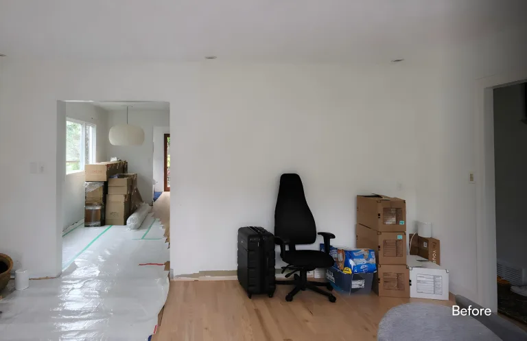 A room prior to renovation with a plain white wall, stacked moving boxes, a black desk chair, a suitcase, and floor protection. — before